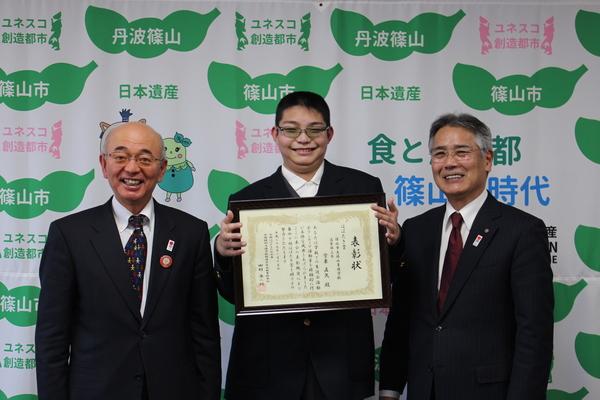 堂東 直矢さんが表彰状を持って、両端には市長と男性が立って笑顔で記念写真