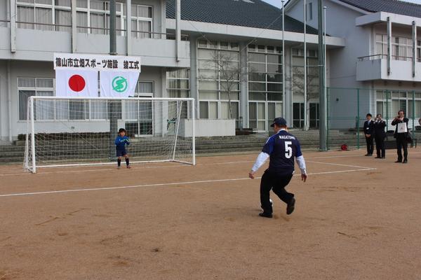 ゴールにはゴールキーパーの選手が構えており、紺色で背番号5のユニホームを着た男性が、ゴールを狙おうとする瞬間の写真