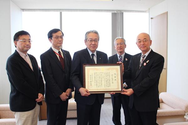 高梨さんが額縁に入った感謝状を手に持って見せており、市長と大国さんと長沼さんと関係者が一緒に写っている写真