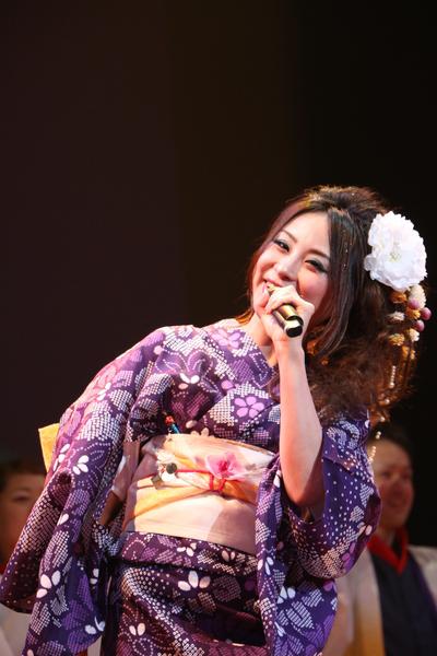 白石 乃梨さんが髪に白い大きな花飾りをつけ紫色の浴衣姿で身体を横にかしげて笑顔で歌っている写真