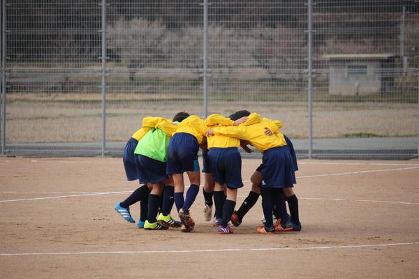 グラウンドで、黄色いユニホームを着たサッカー選手の児童達が円陣を組んでいる様子の写真