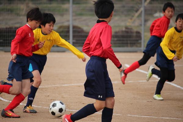黄色いユニホームと赤色のユニホームを着た選手がサッカーボールを競り合って追いかけている様子の写真