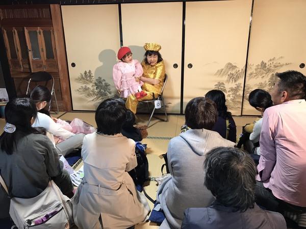 金色の衣装を着た女性が椅子に座り、赤の帽子とピンクの洋服を着た人形を手に人形劇をしており、その話を聞いている方々の写真