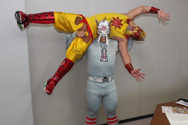 銀色のマスクをかぶり、むねに通天閣のイラストが絵が画れた衣装を着た怪獣ニューワールドが黄色い冠のついたマスクをかぶり、胸に太陽のようなイラストのついた衣装を着ているタコヤキ―ダーを肩の上に持ち上げて、タコヤキ―ダーが両手両足を広げて叫んでいる様子の写真