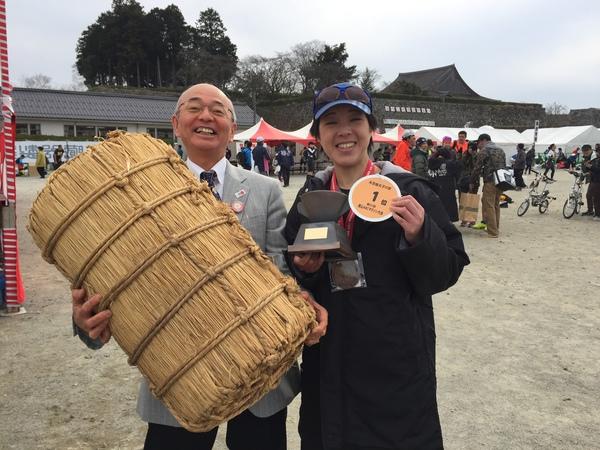 米俵を持った市長と、その横に1位と書かれたメダルと優勝カップを持った女性が笑顔で笑っている写真