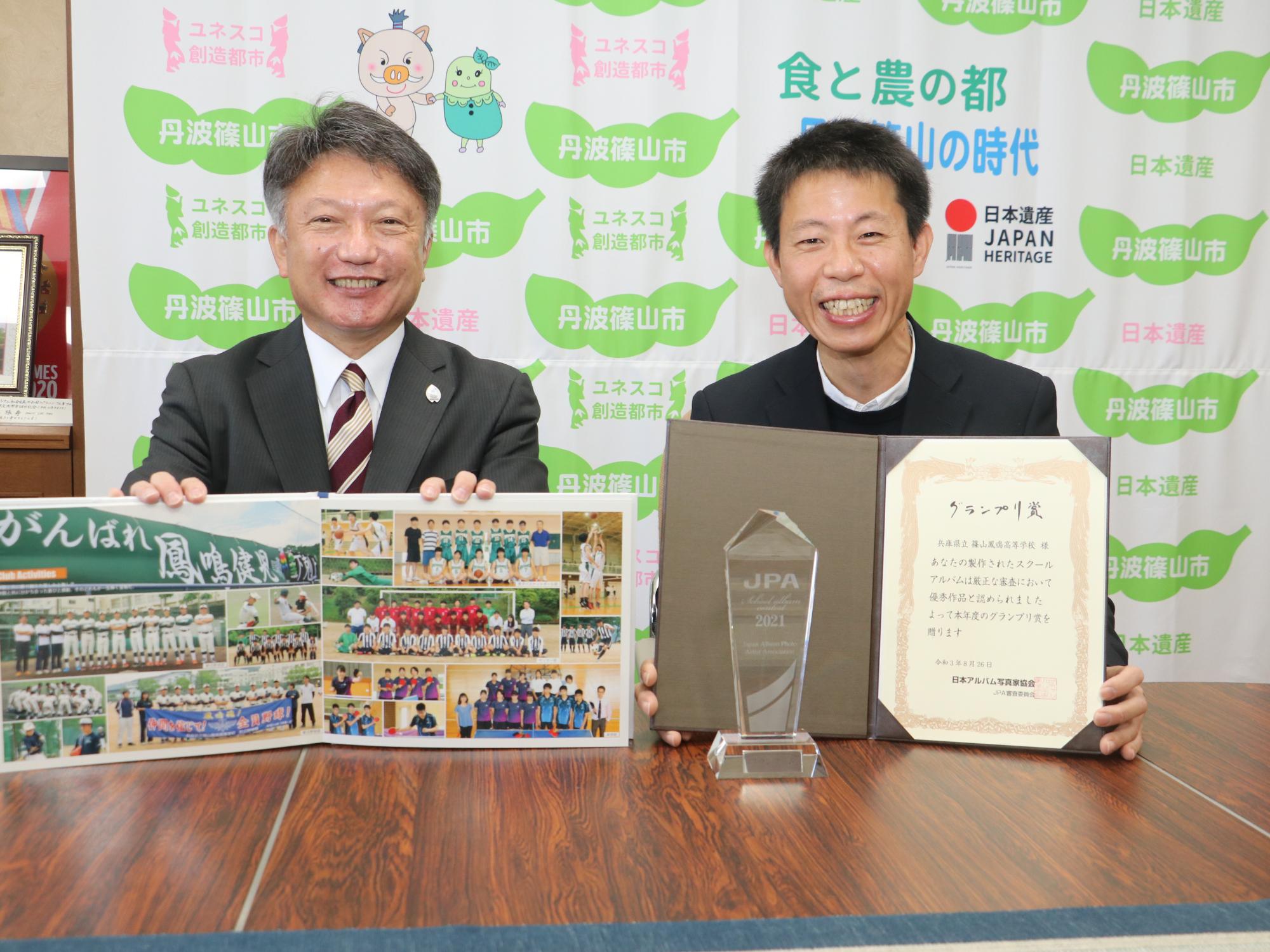 卒業アルバムのページを広げて男性が持って、その隣にグランプリ賞の表彰状を持った受賞者との記念撮影