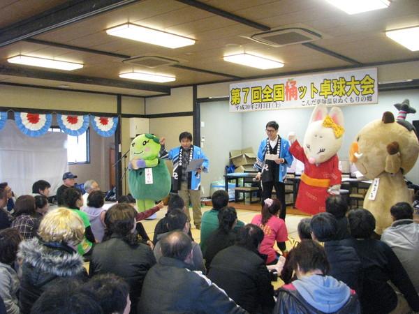 第7回全国桶ット卓球大会の横断幕の前に大会参加者が集まって畳に座っており、参加者の前で、青い法被を着た男性2人がマイクを持って司会進行をしており、きつねの着ぐるみを着た人形と、まめりん、まるいのが写っている写真