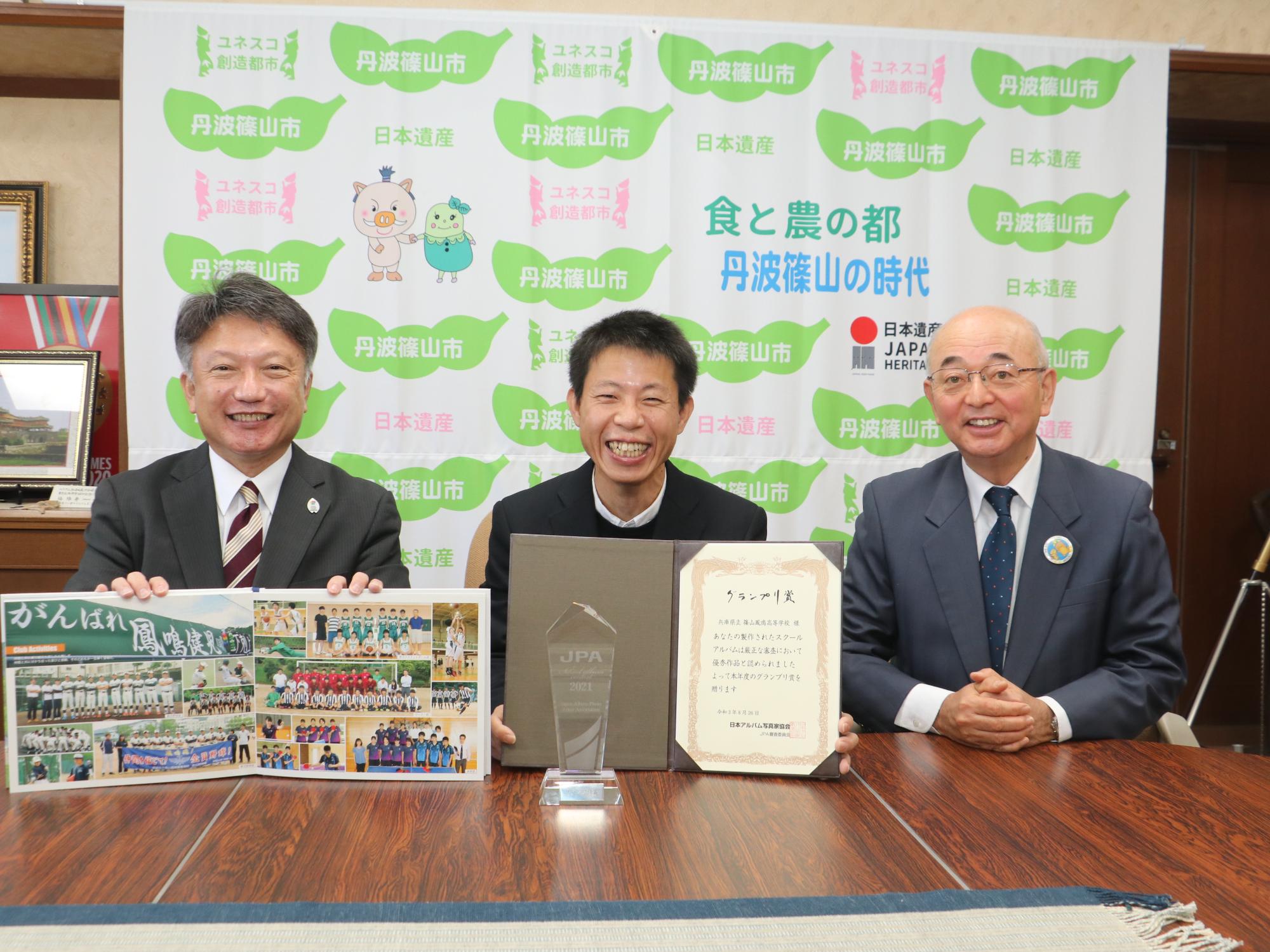 卒業アルバムのページを広げて男性が持って、その隣にグランプリ賞の表彰状を持った受賞者、市長との記念撮影