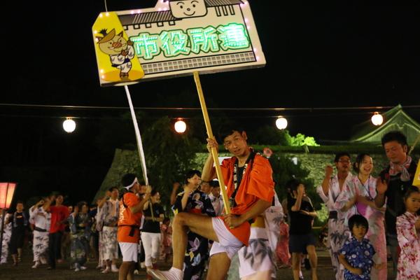 「市役所連」のプラカードを持ったオレンジの法被姿の男性職員を先頭に浴衣姿で踊る人々の写真