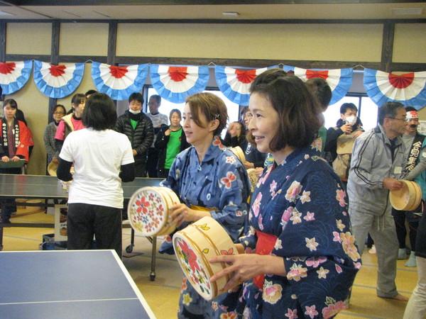 紺色の浴衣を着た女性2人が卓球台の前に立っており、桶を持って構えており、桶の裏に綺麗な色とりどりの花模様が描かれている写真