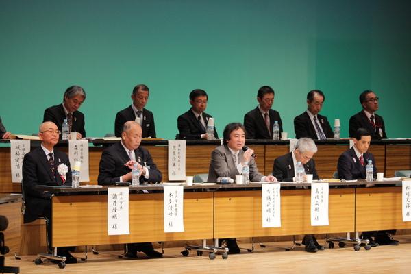 舞台ではスーツを着た男性たちが席に座って、その中の男性がマイクを手に持ち話をしている写真