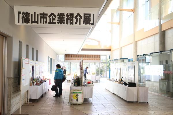 「篠山市企業紹介展」の看板が下がっており、その奥に各企業の製品が机の上に並べられ、パネルで紹介されているブースが設けられている写真