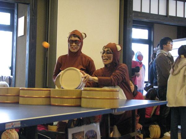 タヌキのコスチュームをしている2人が卓球台の前に立っており、右側1人が桶で飛んできたボールを打とうと構えている様子の写真