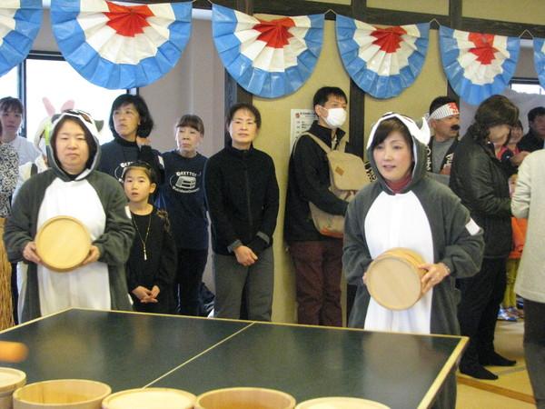 お腹が白く全体がグレーの動物の被り物の衣装を着た2人が卓球台の前に桶を持って構えており、その後ろには沢山の人が立っており、試合を見守っている様子の写真