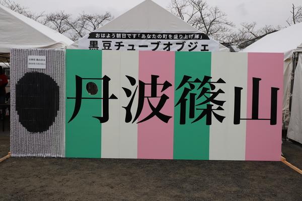 左端に黒い玉のようなもので丸く描かれた物と丹波篠山と書かれた文字の看板が設置されている写真