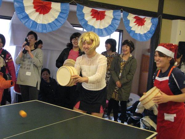 金髪に白いブラウスと黒いミニスカートを着た男性と、サンタの帽子と赤いエプロンを付けた女性が、卓球台の前に桶を持って立っており、飛んで来たボールを打とうと構えている様子の写真
