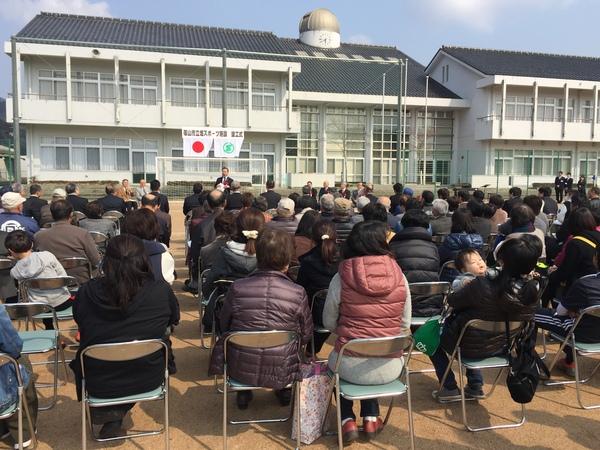 グラウンドに並べられている沢山の椅子に出席者が座っており、前方で黒いスーツを着た男性の話を聞いている様子を写している写真