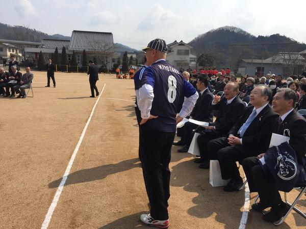市長が背番8の紺色のユニホームを着ている様子を背後から写しており、右側には沢山の出席者が椅子に座っている様子の写真