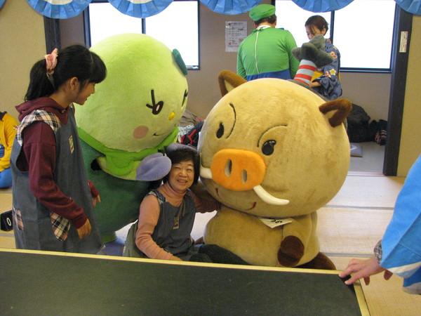 まめりんとまるいのが、座り込んでいるエプロンを付けたショートカットの女性の頭と背中をやさしく触っており、その様子を同じチームのエプロンを付けて髪を一つに結んでいる女性が見ている様子の写真