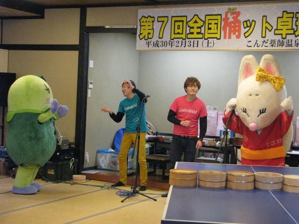 スタンドマイクの前で、きつねの着ぐるみを着た人形が右手でマイクを握って左手を上げており、青いティーシャツを着た男性とまめりんが向かい合って踊っており、ピンク色のティーシャツを着た男性がきつねの着ぐるみを着た人形を見ている様子の写真