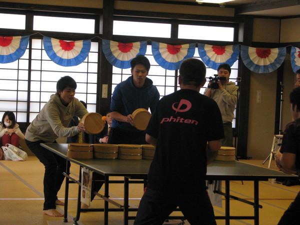 黒色で「Phiten」の文字がバックプリントされたティーシャツを着た男性チームとグレーのパーカーと青色のパーカーを着た男性チームが真剣な表情で試合をしており、その様子をテレビカメラが撮影している様子の写真