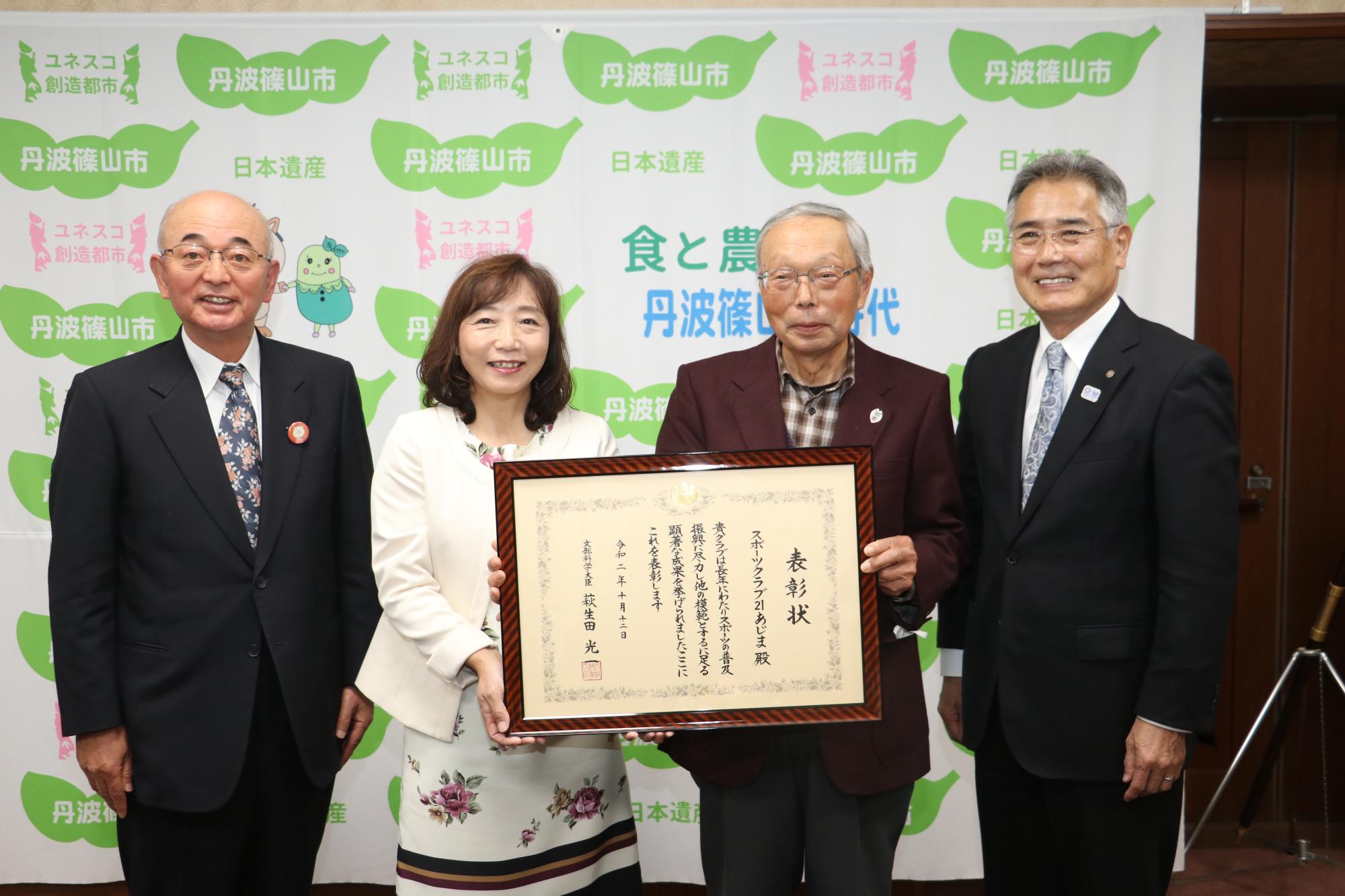 額に入った表彰状を手に持っているスポーツクラブ21あじまの方、その隣に酒井市長、前川教育長が立っている