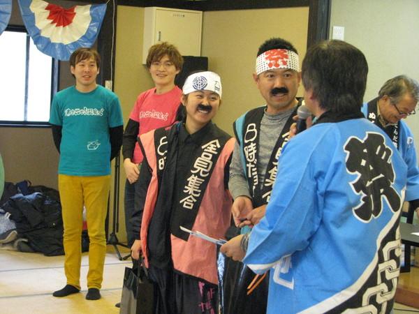 頭には豆絞りを巻き、8時だヨ 全員集合と書かれている法被を着て、鼻ひげを付けている男性と女性にマイクを持った男性がインタビューをしている様子の写真