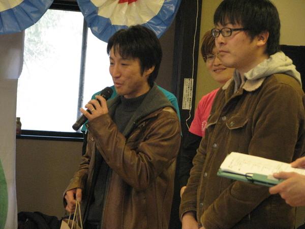 茶色の革ジャンを着た男性がマイクを持って話をしており、隣の男性は話を聞いている様子の写真