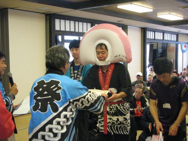 かまぼこの被り物をしている男性が、大会参加者の前で表彰を受け、「祭」とかかれている青い法被を着た男性から、賞状を受け取っている様子の写真