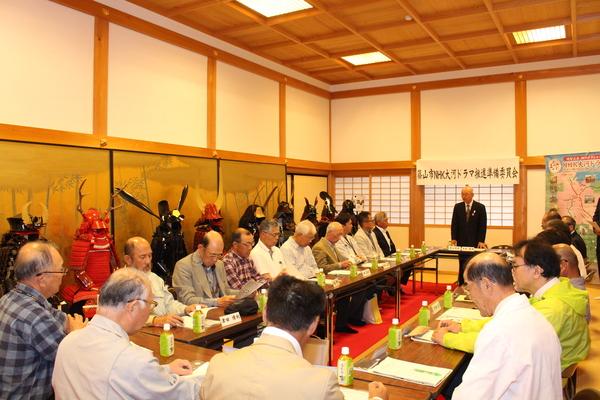 篠山市NHK大河ドラマ推進準備委員会にて市長が参加者の前で立ち上がって話をしていおり、参加者は椅子に座って机の上の資料に目を向けて話を聞いており、左側には沢山の武将の甲冑が飾られている様子の写真