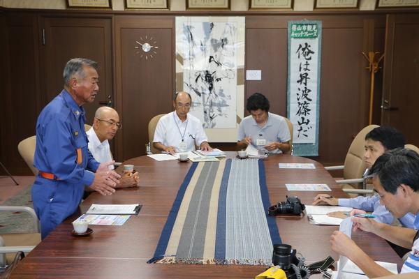 消防団員が立って話をされていて、テーブルにカメラを置いてメモを取っている方の写真