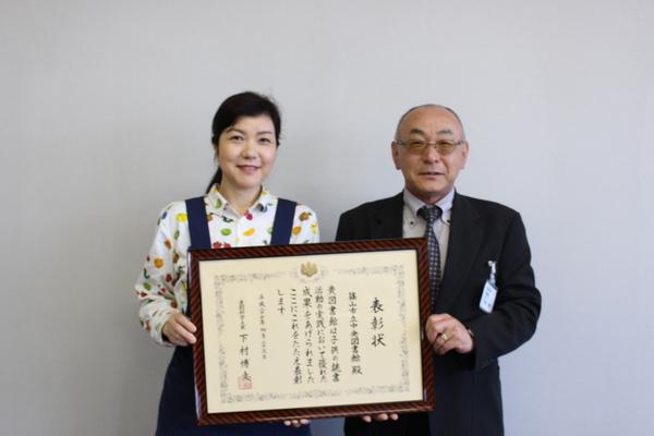 図書館の赤井毅彦館長と司書の松下順子さんが、笑顔で額入りの表彰状を持っている写真