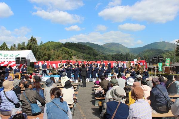 青空の下、大山荘の里市民農園収穫祭のステージで行われている丹南中学のブラスバンドの演奏を聞いている人々の写真
