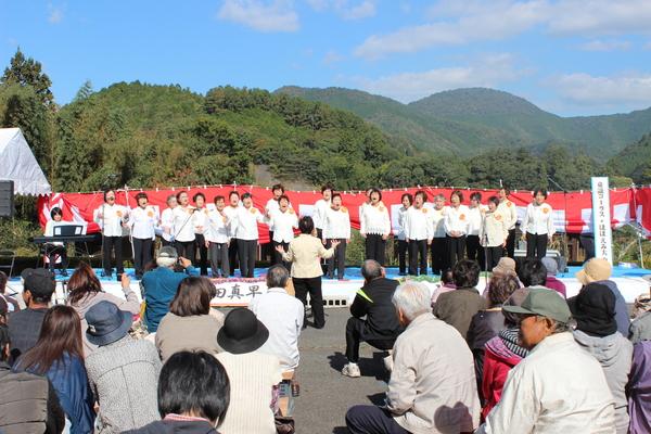 青空の下、大山荘の里市民農園収穫祭のステージで女性コーラスグループが指揮に合わせて歌を歌っているのを見ている人々の写真