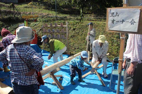 里山オーナー会の会場で男の子がのこぎりで木材を切っている様子を見ている大人たちの写真