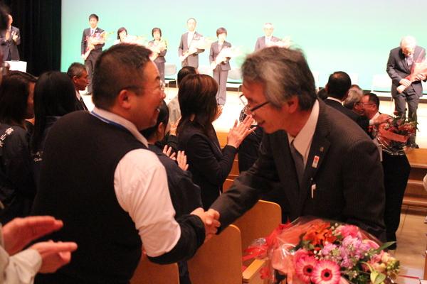 退場する退職者の男性が会場にいる1人の男性と握手を交わしている写真