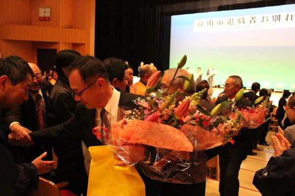 花束を抱えた退職者の男性が拍手で送られている写真
