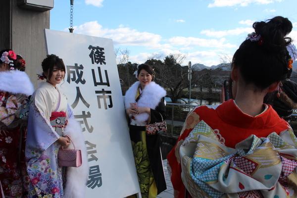 「篠山市成人式会場」と書かれた看板を間に挟んで、振袖を着た女性二人が記念写真を撮っている様子を後方から写している写真