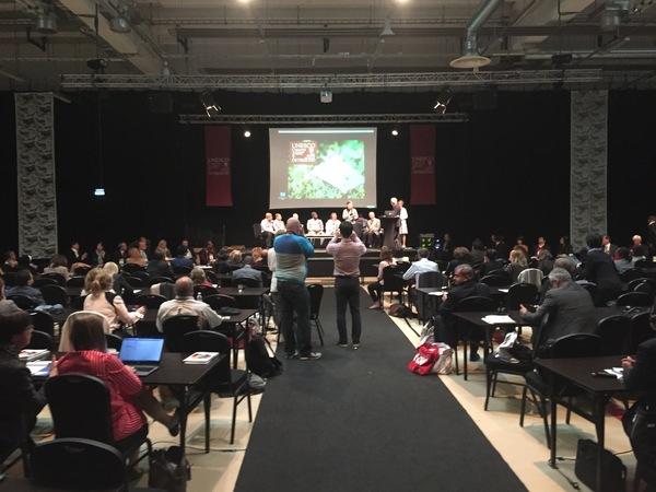 舞台の上のスクリーンに映像が映っており、その前でマイクを持った方が発表している、その発表内容を海外の方が真剣に聞いている写真