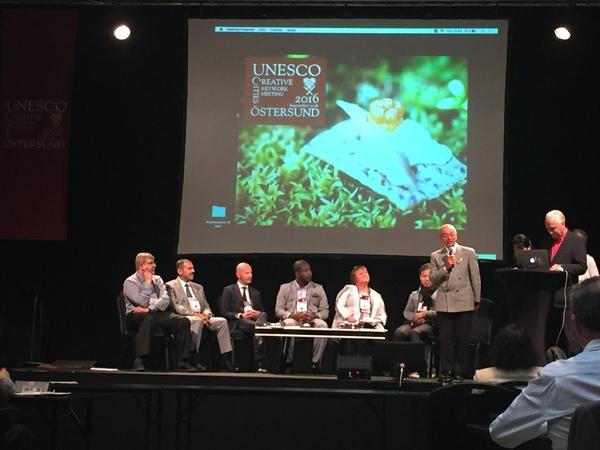 舞台にスクリーン映像が映し出されており、その舞台の上で外国の方が椅子に座り、市長の話を聞いている写真