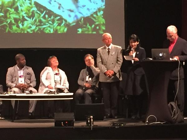 舞台の上のスクリーンに映像が映っており、そのスクリーンの前で市長が女性と一緒に立ち、女性が紙を見ながらみんなの前でお話ししている写真