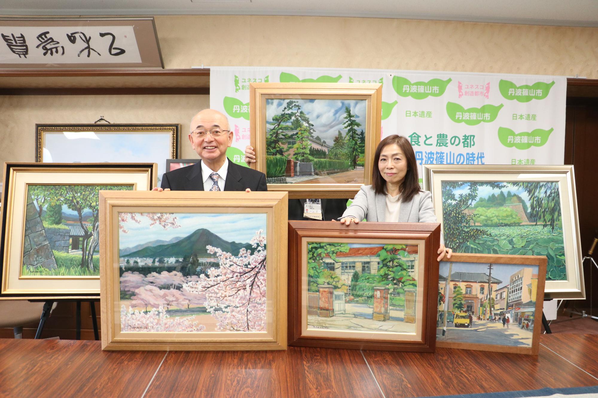 寄贈していただく絵画を机の上に3点、後ろに3点並べて寄贈者の荒川さんと市長の記念撮影