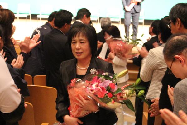 拍手で送られ花束を抱え退場する女性退職者の写真