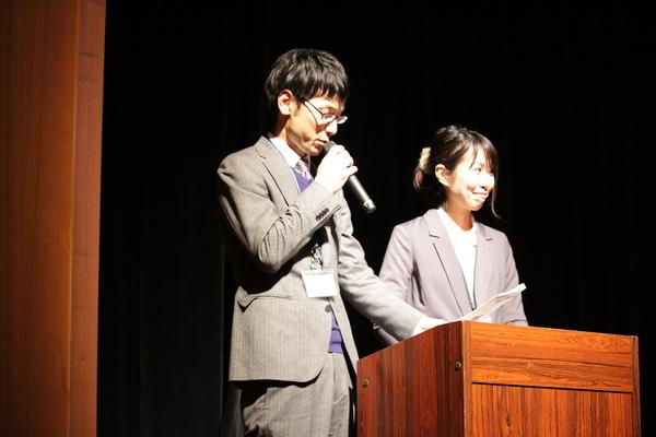 壇上に上がり業務改善の実践事例をマイクで発表している男性職員と女性職員の写真