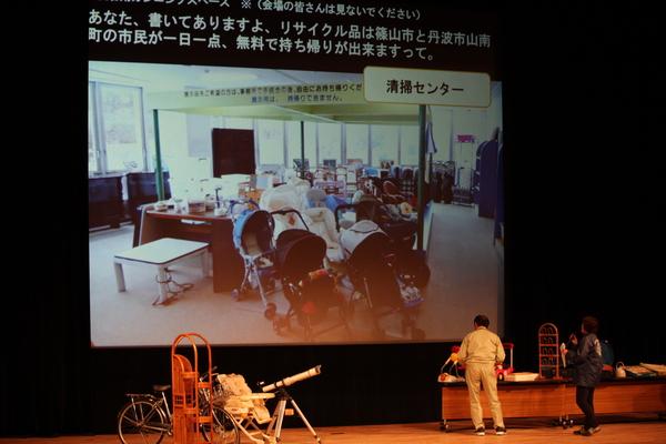 準グランプリ「清掃センターチーム」のプレゼンテーションの様子(壇上には捨てられた家具、自転車、望遠鏡などが置いてあり、スクリーンではリサイクル品展示の様子が映し出されている)