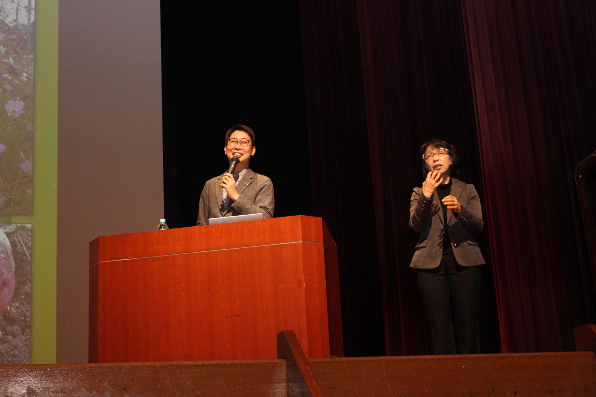 舞台上でマイクを手に持って講演される片平さん。隣には手話通訳者の女性が立っている。