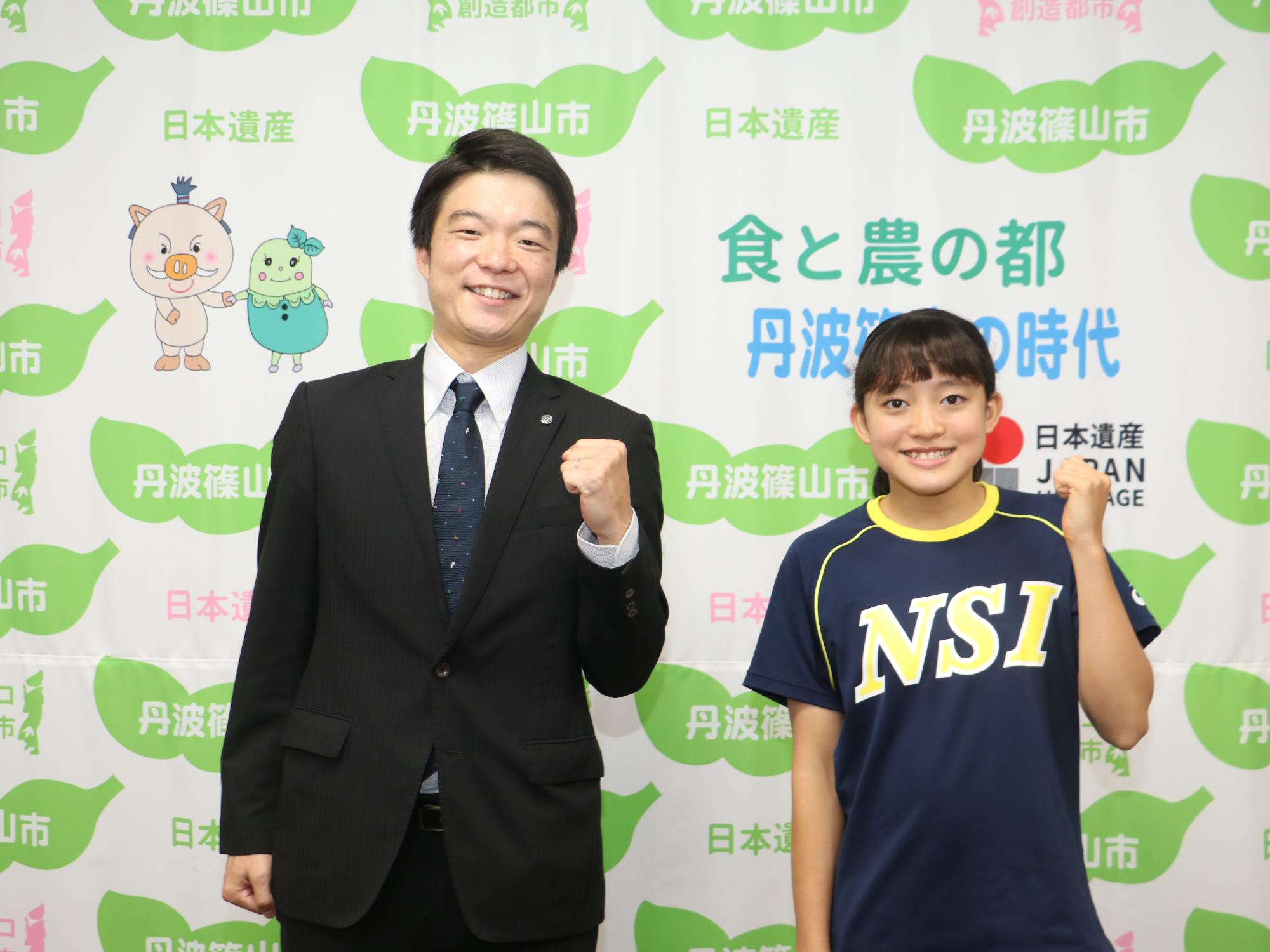 スーツを着た男性と水泳チームのユニフォームを着た女性が左手でガッツポーズをしている