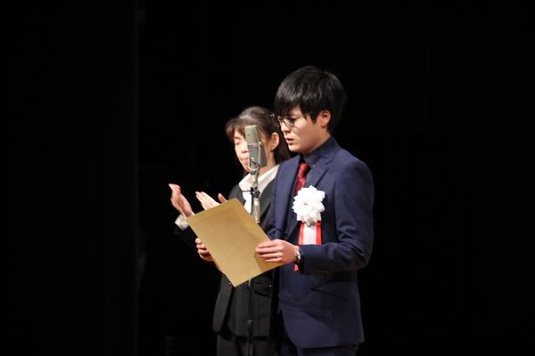 実行委員長の大見君がスタンドマイクの前で話をしており、その左隣りで、通訳の女性が同時通訳をしている様子の写真