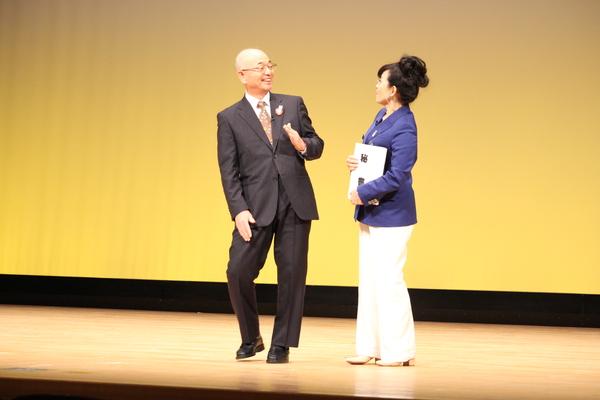 市長が紫色のジャケットを着た秘書役の女性と話をしている寸劇の場面の写真