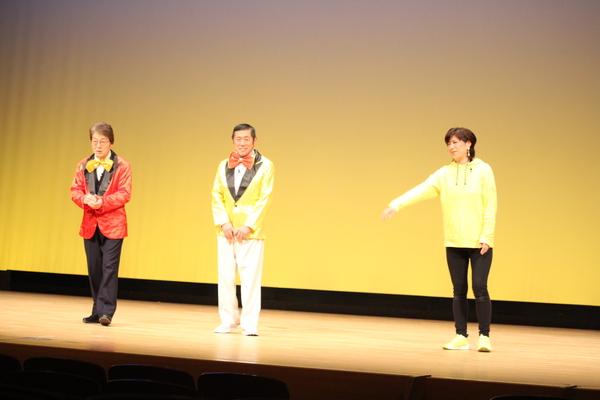 赤いジャケットに金色の蝶ネクタイの男性と、金色のジャケットに赤い蝶ネクタイの男性が立っており、黄色いパーカーを着た女性が右手を前に出している寸劇の場面の写真